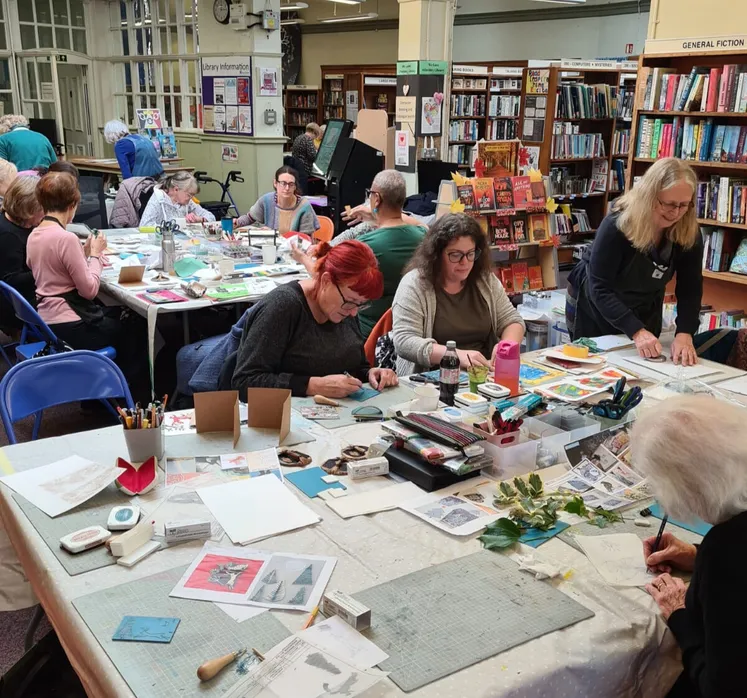 A group of adults gathered around tables in a library, engaged in arts and crafts. They are working with paper, stamps, and drawing tools, surrounded by supplies like scissors, ink pads, and printed designs. Bookshelves filled with books line the background.