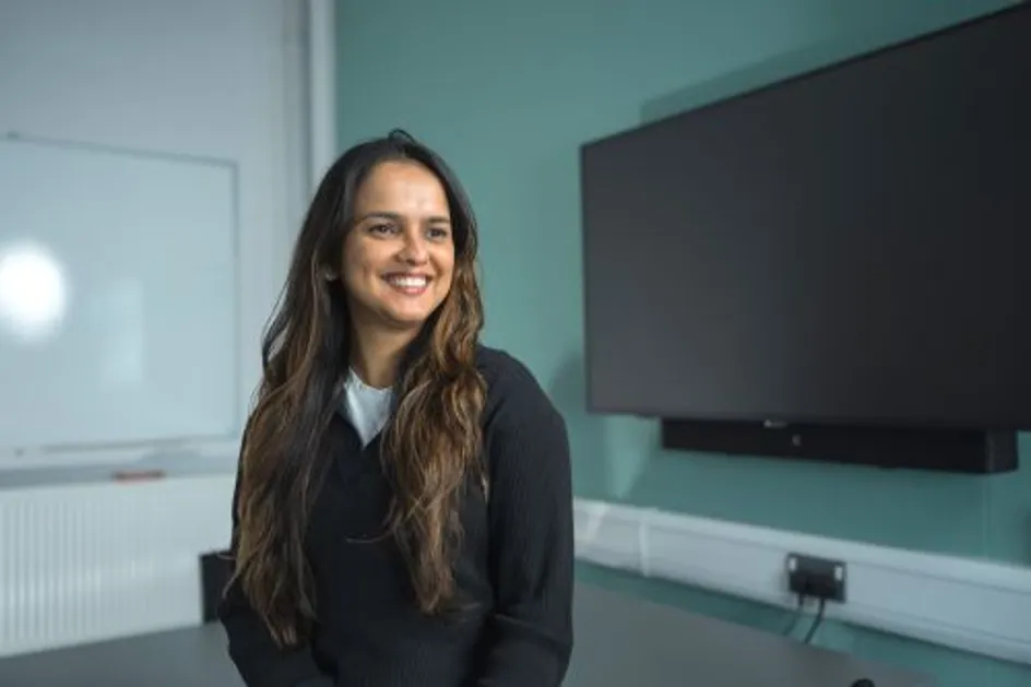 Chamathka is stood in a classroom in front of a large TV screen. She is wearing a black jumper and smiling for the camera.