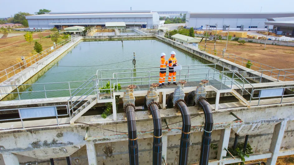 Two men in orange overalls, standing in front of a large wastewater treatment pond