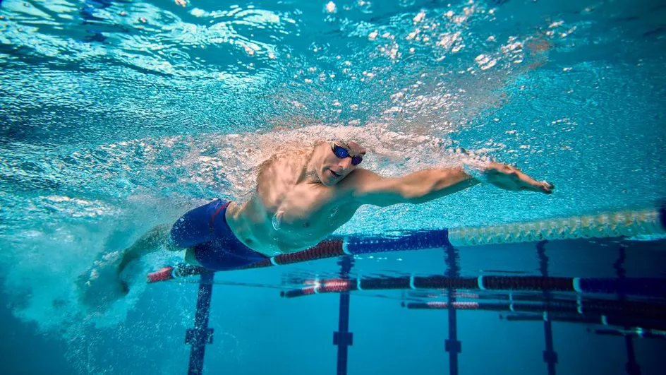 Underwater view of a swimmer performing a freestyle stroke in a swimming pool, wearing goggles and blue swim trunks.