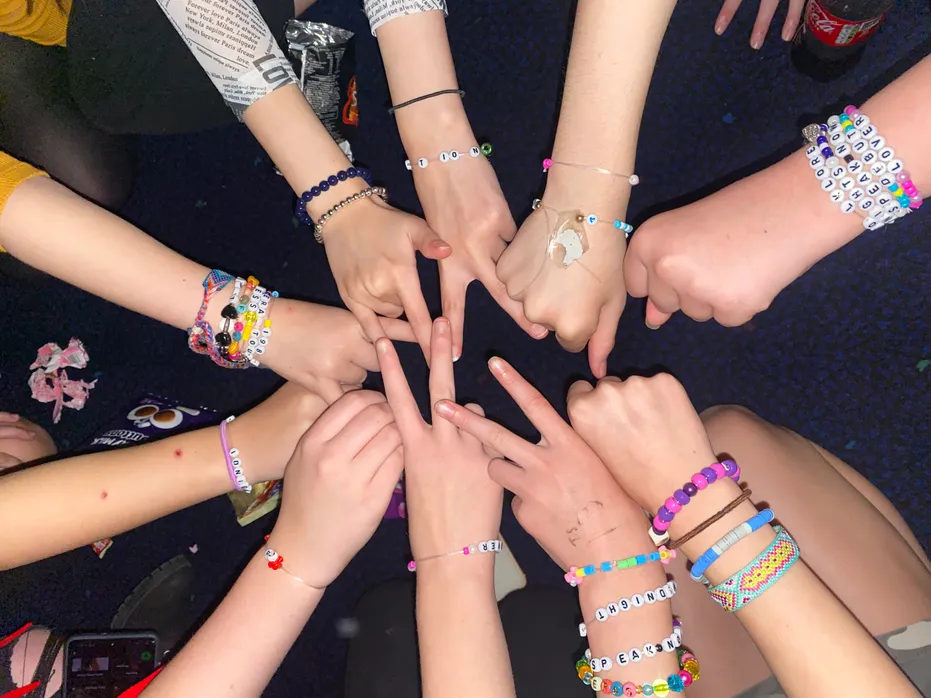 Close-up of eight hands reaching into the centre of a circle, each wearing colourful handmade friendship bracelets and beaded wristbands. Some hands form heart shapes or point towards the middle, creating a sense of connection and shared experience.