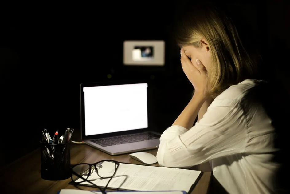 A person sits at a desk in a dark room at night, covering their face with both hands while looking toward a brightly lit laptop screen.