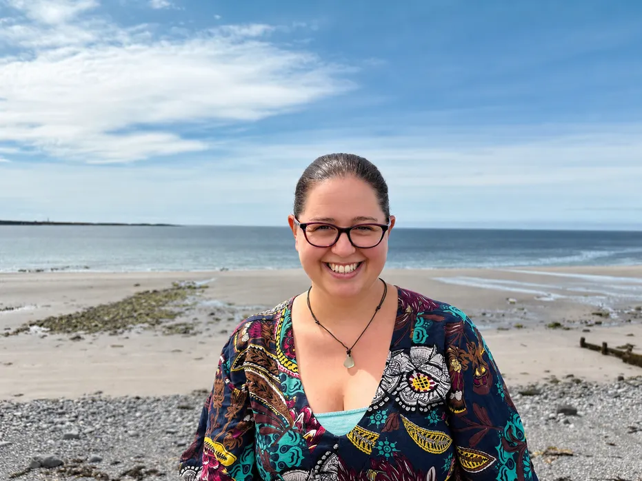 Heather smiles at the camera on a pebble beach with the sea and a blue sky behind her. The shoreline is stretching into the distance.