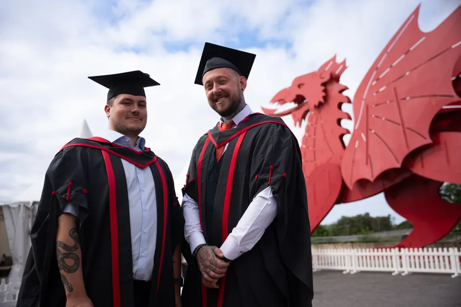 Ross Wills a Connor Jones, yn eu gwisg raddio a’u hetiau graddio, o flaen draig enfawr y tu allan i'r ICC yn Casnewydd, Cymru