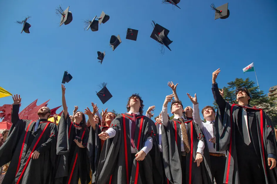 A group of graduates wearing black academic gowns with red trim stand outdoors under a clear blue sky, tossing their mortarboards into the air during a graduation celebration.