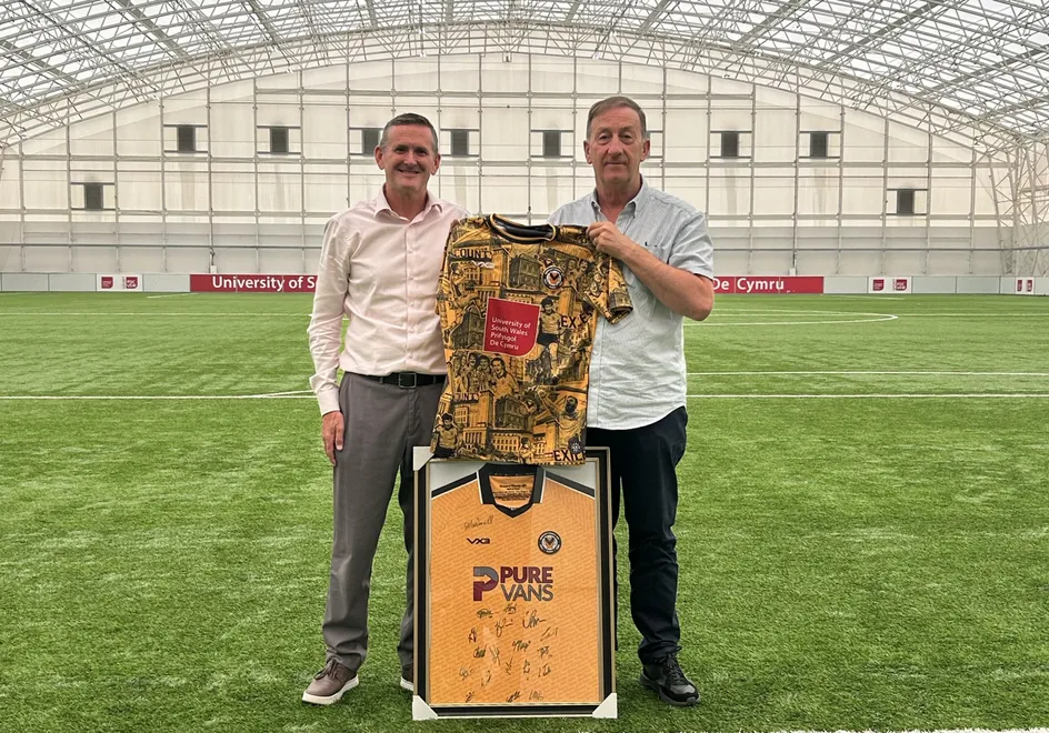 Two people stand on an indoor football pitch beneath a large arched training dome, holding a patterned Newport County football shirt featuring a University of South Wales logo. In front of them is a framed signed Newport County jersey.
