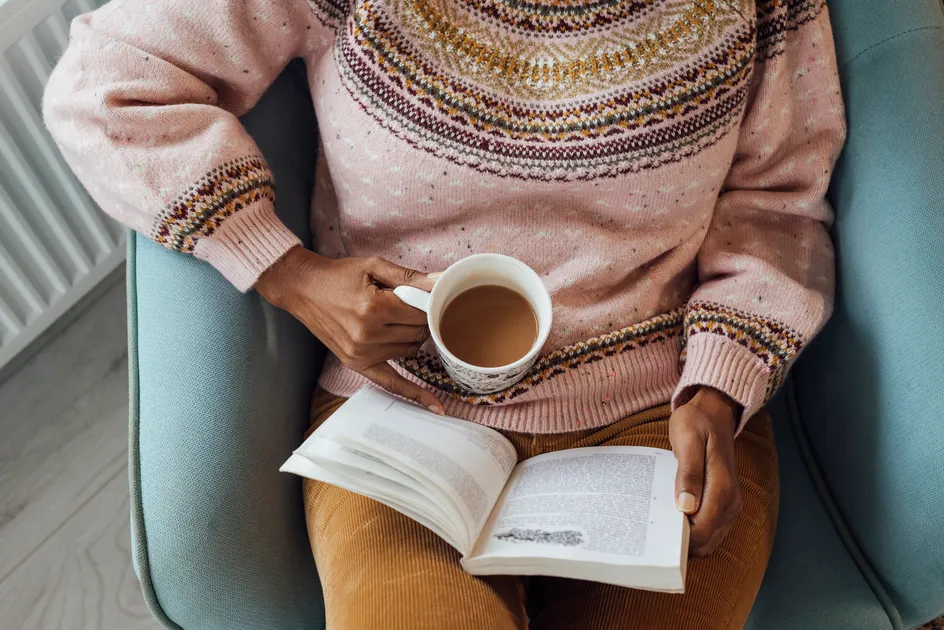 A person is sat in an armchair. They are reading a book and holding a cup of tea.