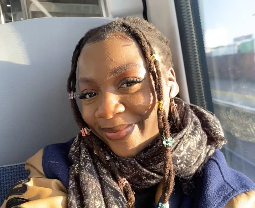 Anodiwa sits by a window on a train, wearing a dark scarf and a blue and tan jacket. Several colourful clips decorate her long braided hair.