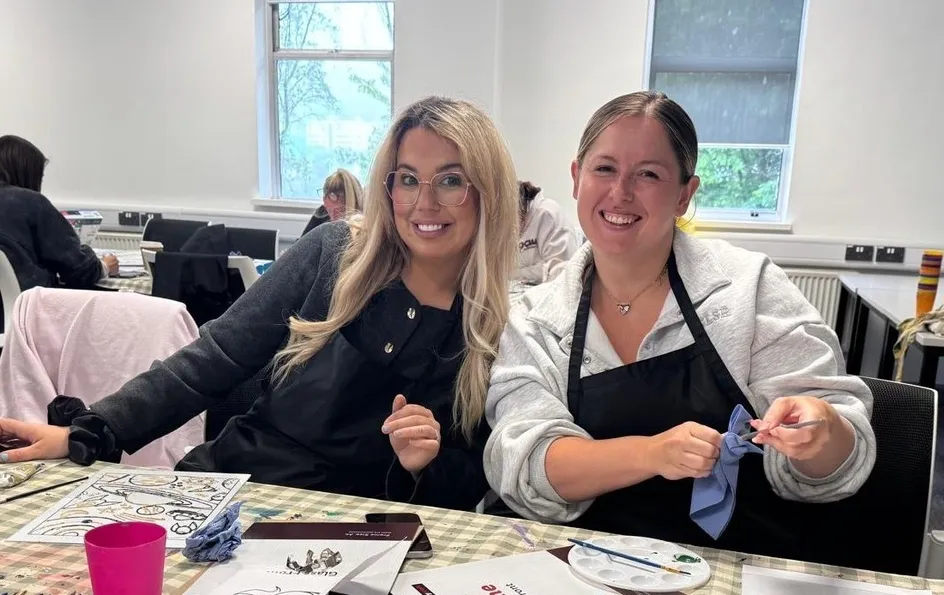 Two students sit together at a table during a wellbeing activity, there are paints and brushes on the table.