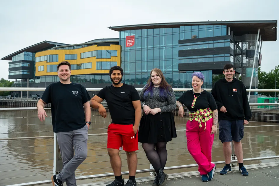 Mae pump o bobl yn sefyll ar hyd llwybr ar lan afon o flaen Campws Casnewydd Prifysgol De Cymru. Maen nhw'n ymgynnull wrth rŵg fetel wrth ymyl y dŵr, gyda'r rhannau melyn a du nodedig o'r adeilad yn weladwy yn y cefndir. Mae un person yn gwisgo siorts coch llachar ac mae un arall yn gwisgo trenis pinc llachar.