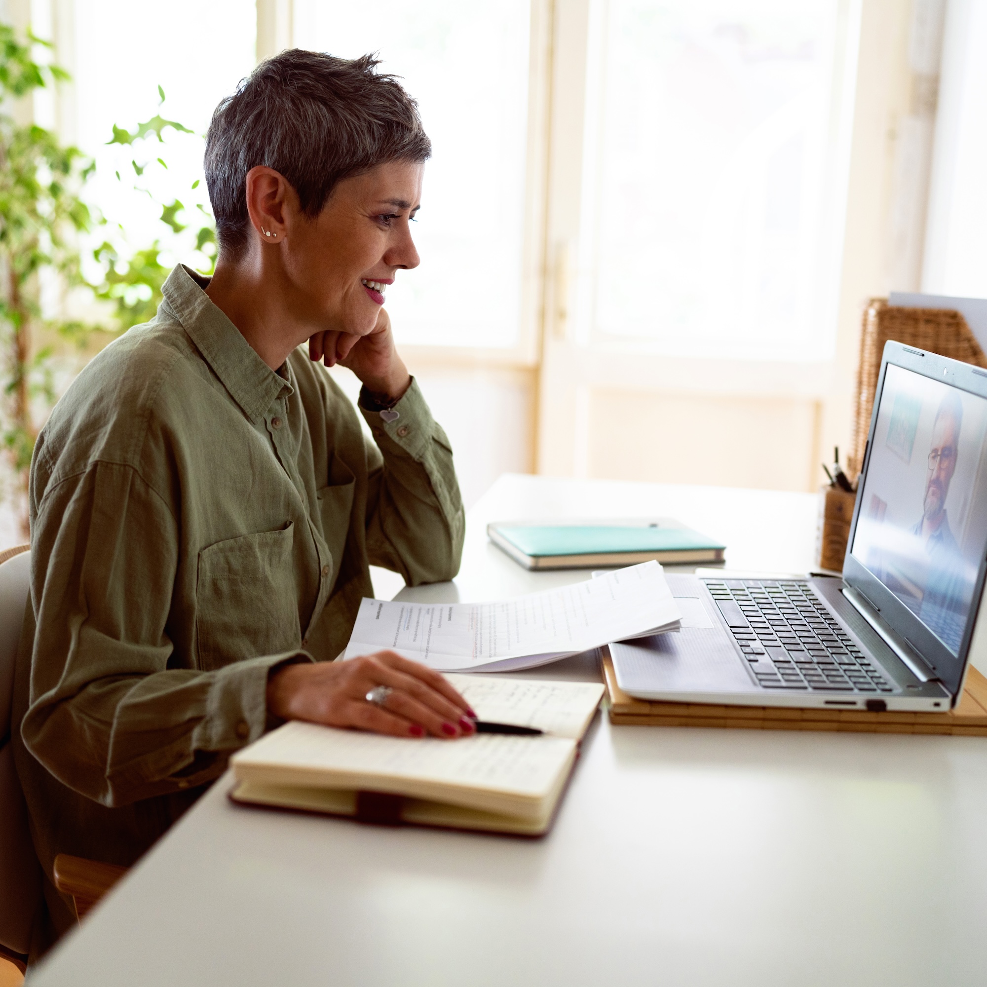 A mature student sits at a desk with their laptop, notebooks and pens in their home, following an online course on the laptop