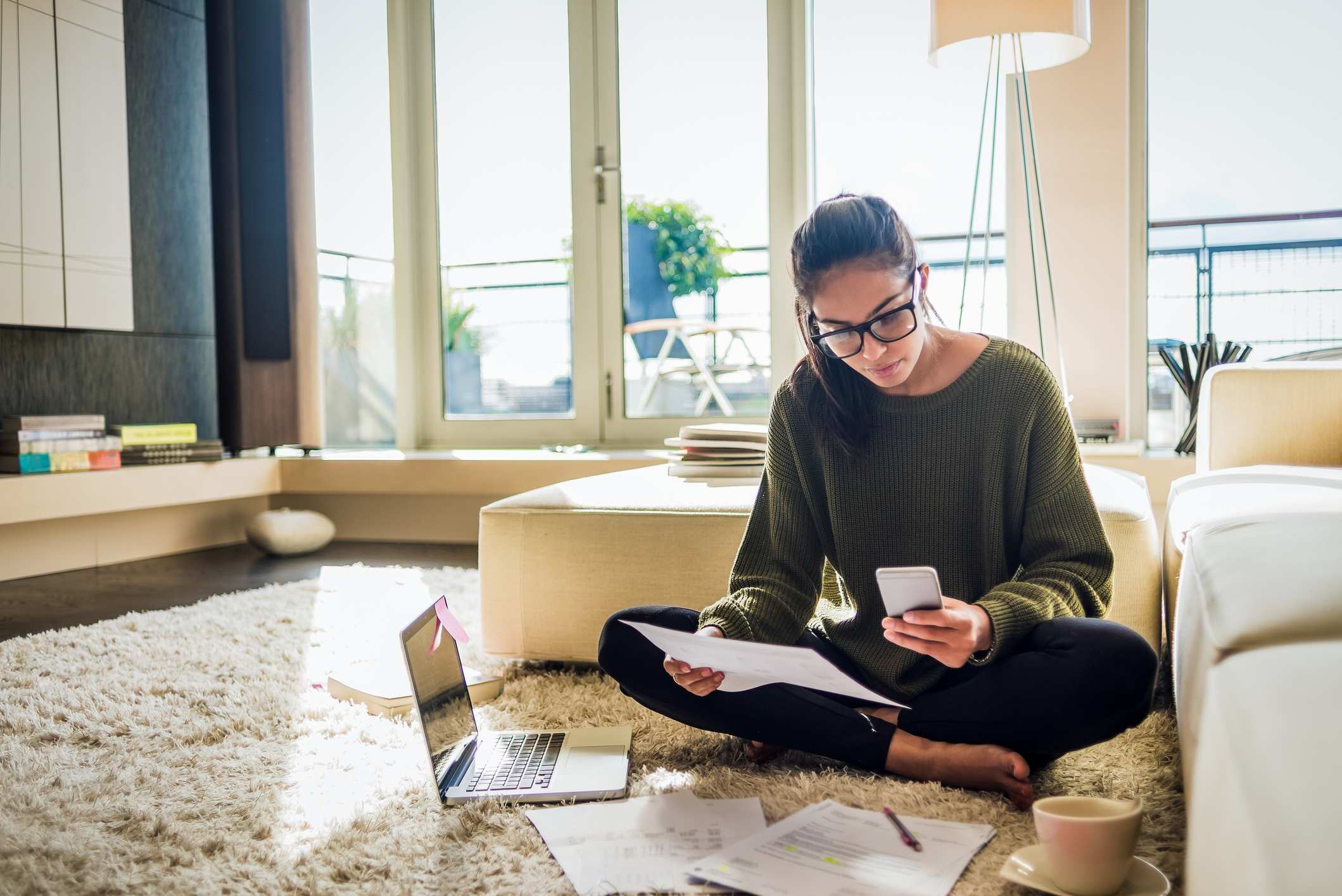 A person sat on the floor surrounded by papers and a laptop, using a calculator.