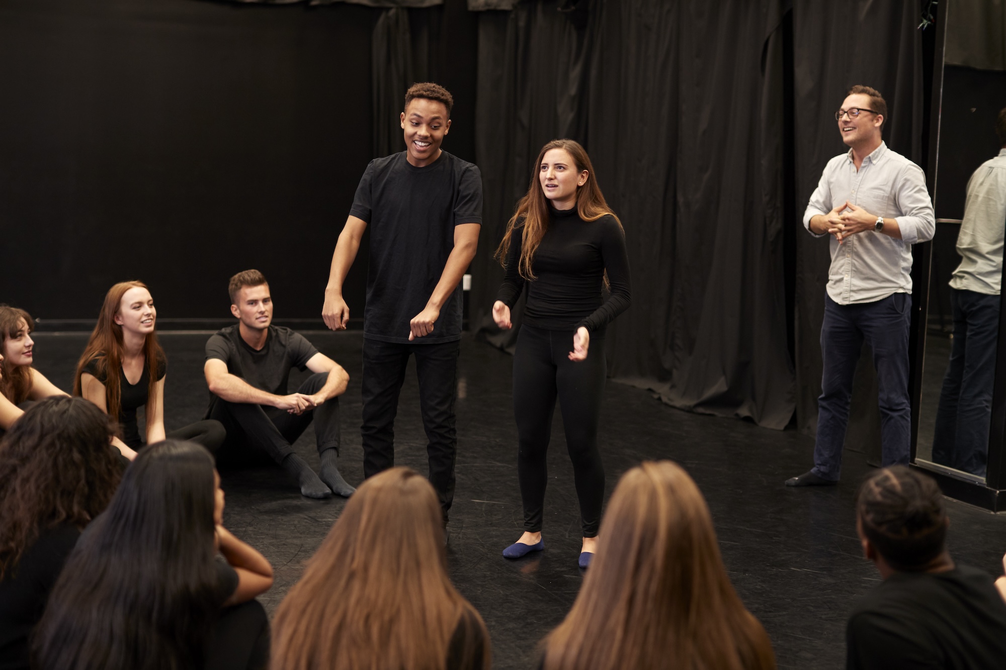 An academic watches two drama students performing for their classmates in a black drama rehearsal studio