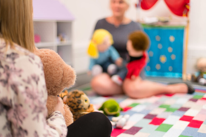 A woman is performing a puppet show for a young girl holding a teddy.