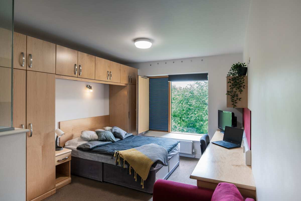 bedroom in mountain halls studio flat showing a double bed with bedside table, wardrobe and cupboards, desk with laptop and TV, desk chair and window