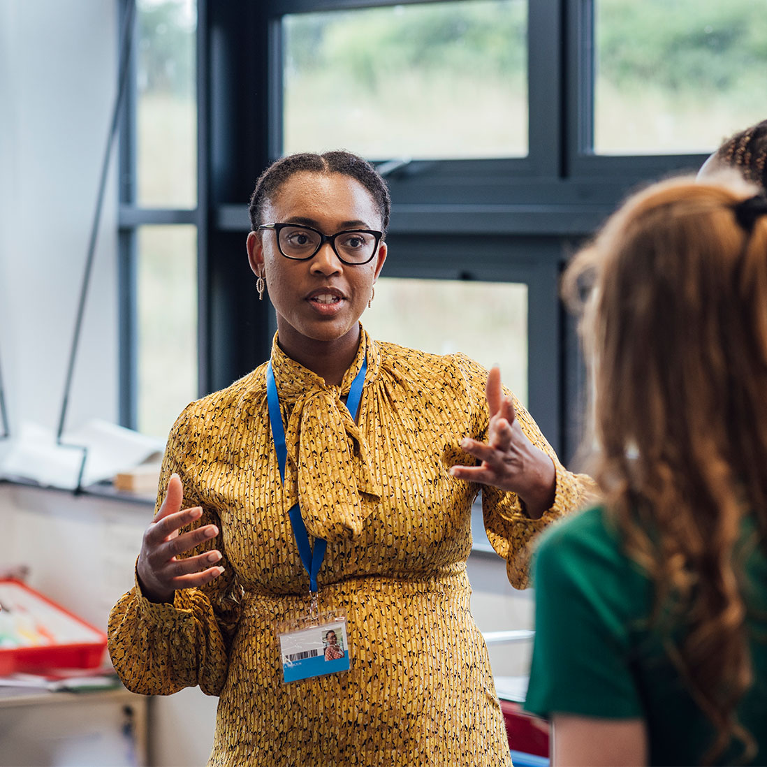 An academic wearing a blue lanyard talks while animating points with their hands to a couple of students while stood in front of windows in a classroom