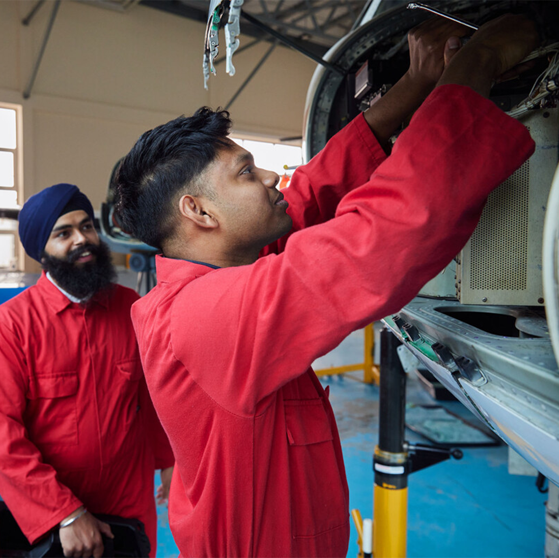 A student in red overalls repairs part of a small aircraft as another student watches