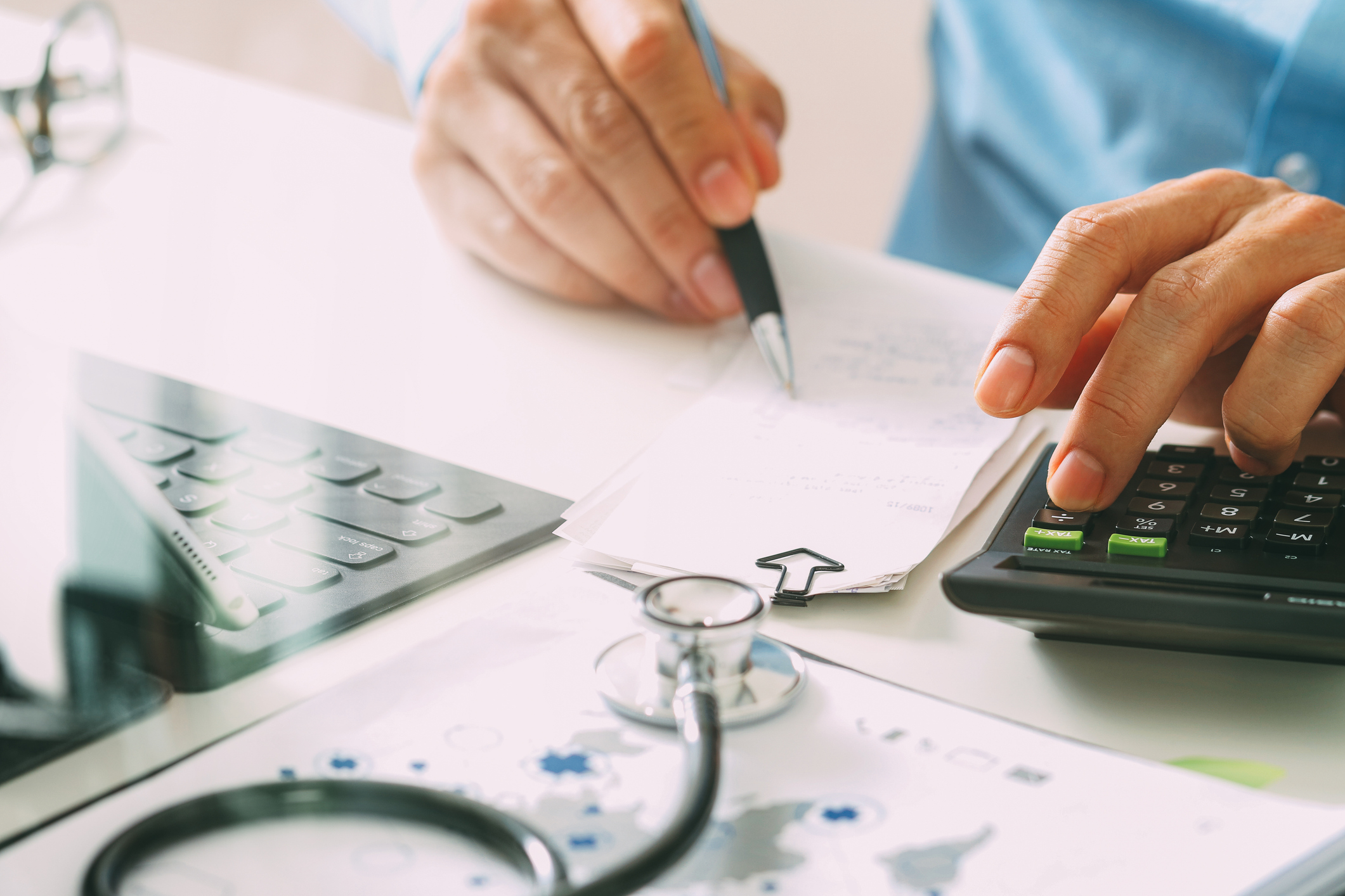 The hands of a doctor writing a note and using a calculator at a desk with a laptop and a stethoscope