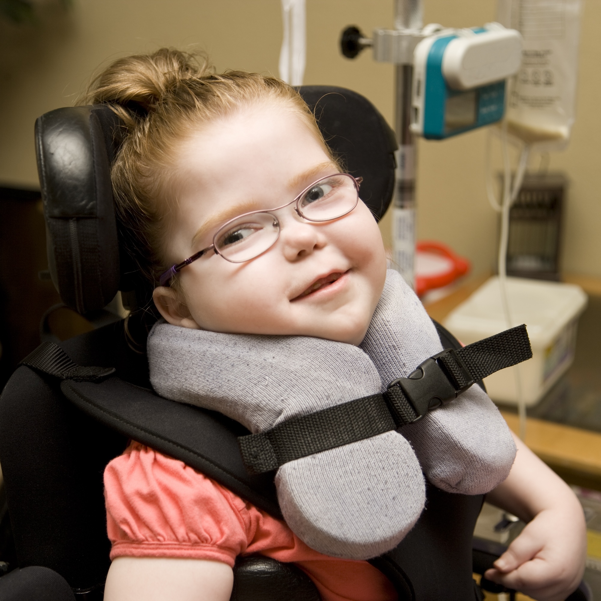 Little girl with cerebral palsy smiles at camera while sitting in her wheelchair at home with other medical equipment in the background