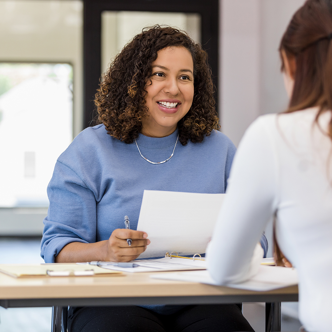 A professional smiles and chats to another professional across a desk while holding an A4 sheet of notes