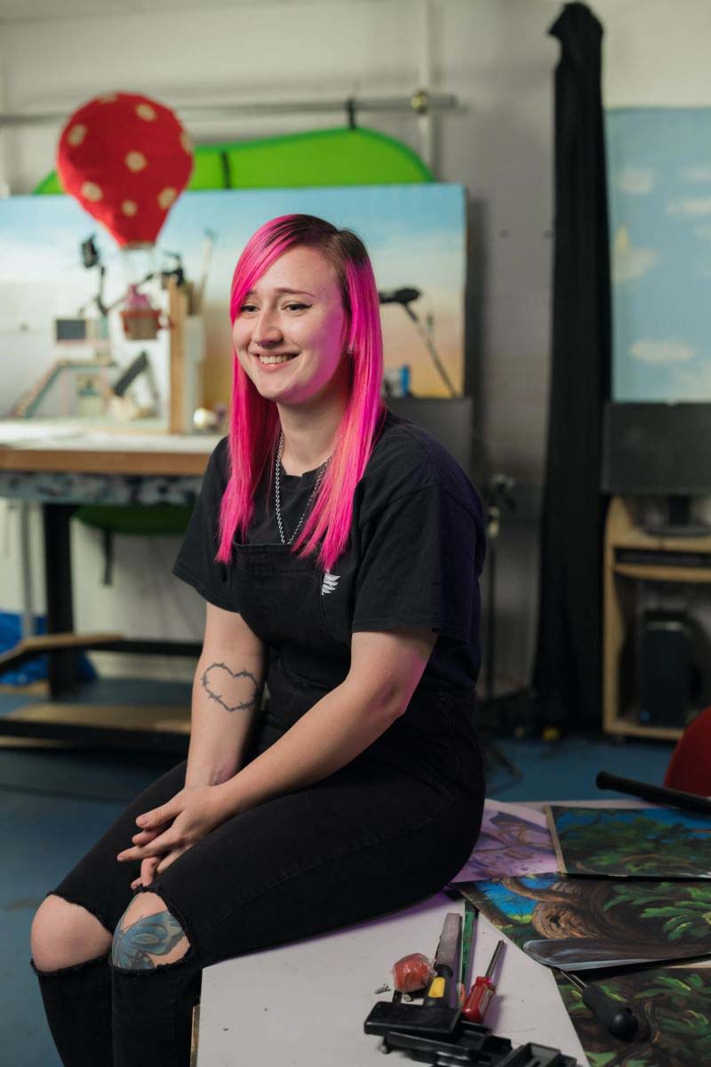 A profile shot of a woman with pink hair sitting at a table surrounded by various art supplies.
