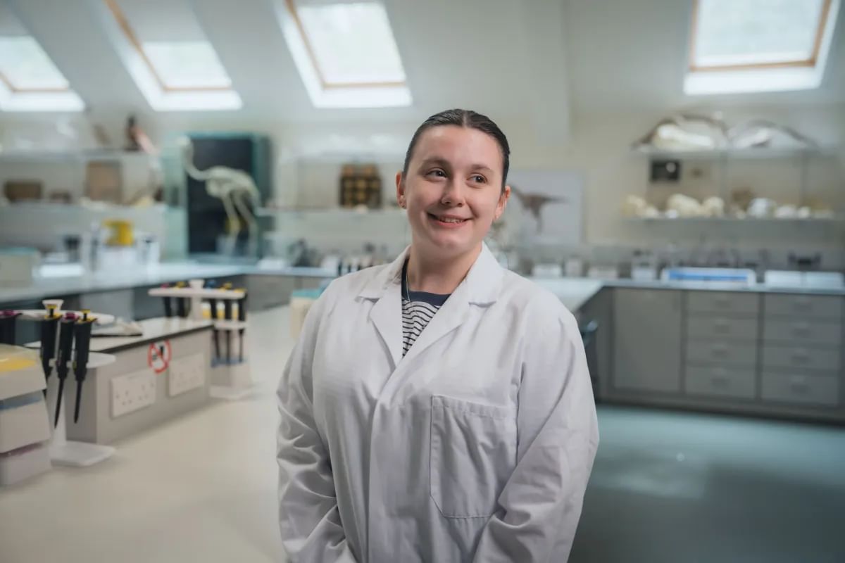 A biological science student wearing a white lab coat, smiling at the camera in a bright, modern laboratory filled with scientific equipment and models.