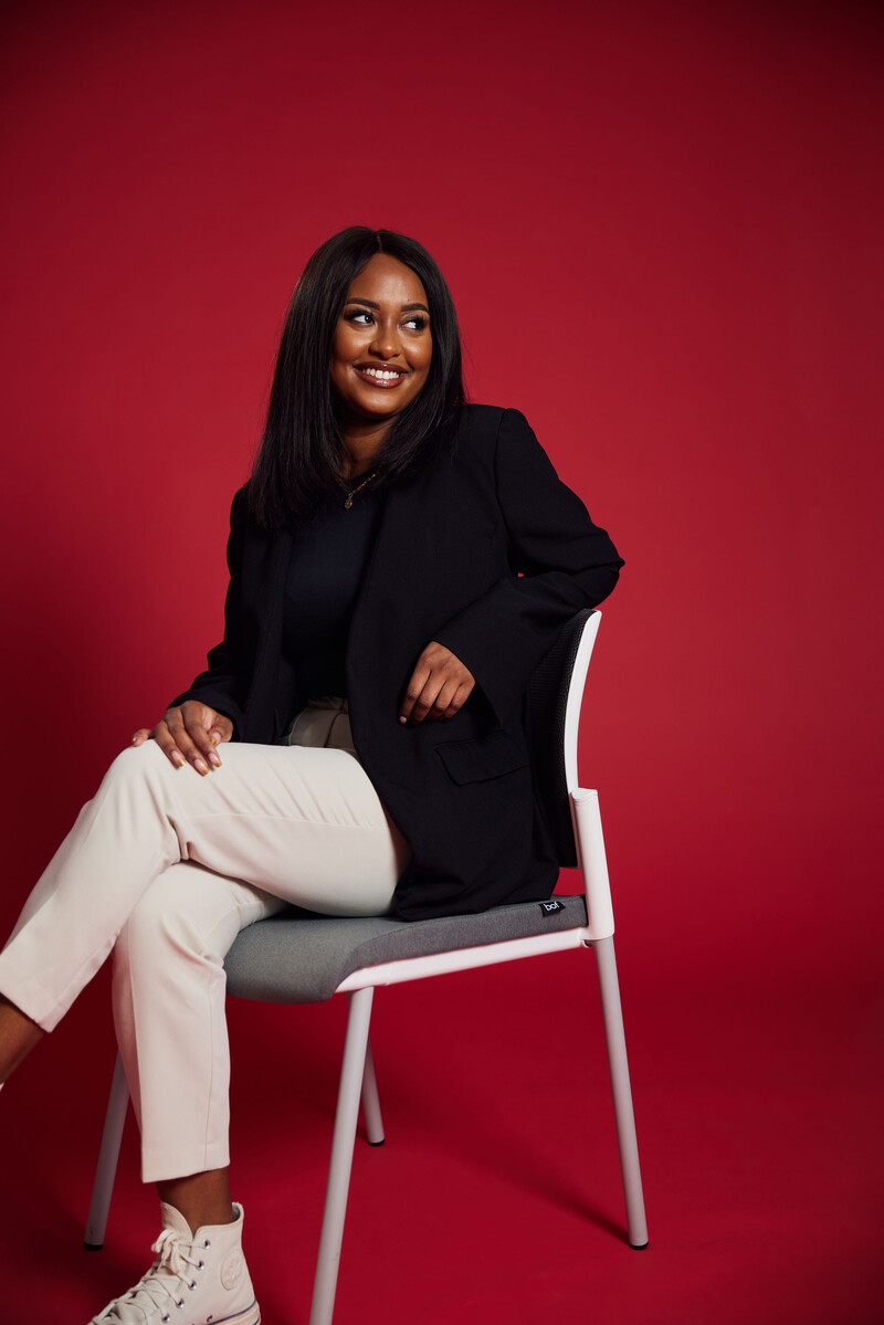 Student Nadifa Saleh sits with their legs crossed on a chair against a red backdrop