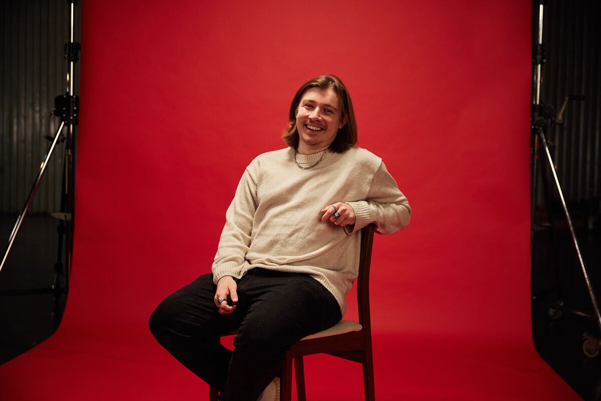 Cyber Security alumni, Sam, sat in front of red backdrop looking directly in to the camera