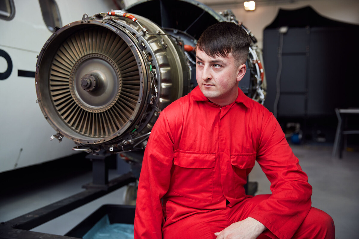 Aircraft Maintenance Engineering student Matthew Dyke poses in red overalls in front of the engine on a small plane in an aerospace workshop