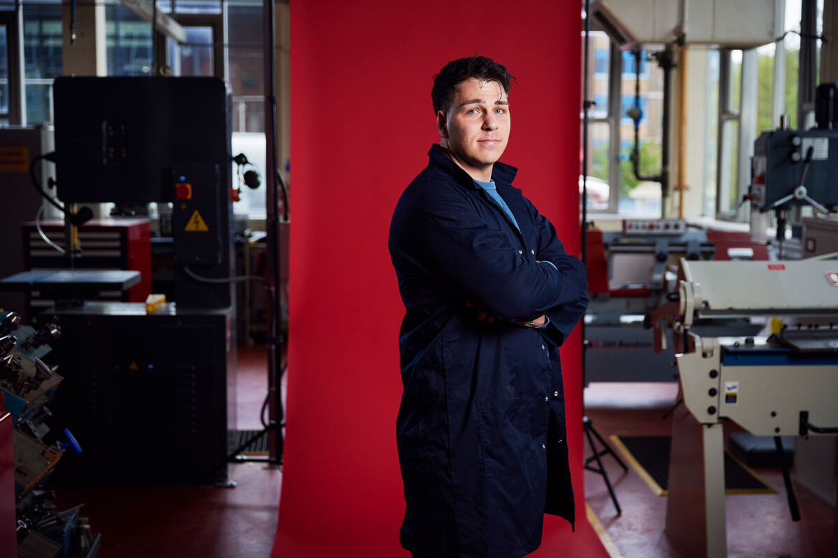 Mechanical Engineering student Santino Petrosillo stands with their arms crossed while wearing a navy engineering coat in front of a red backdrop surrounded by industry equipment in an engineering workshop