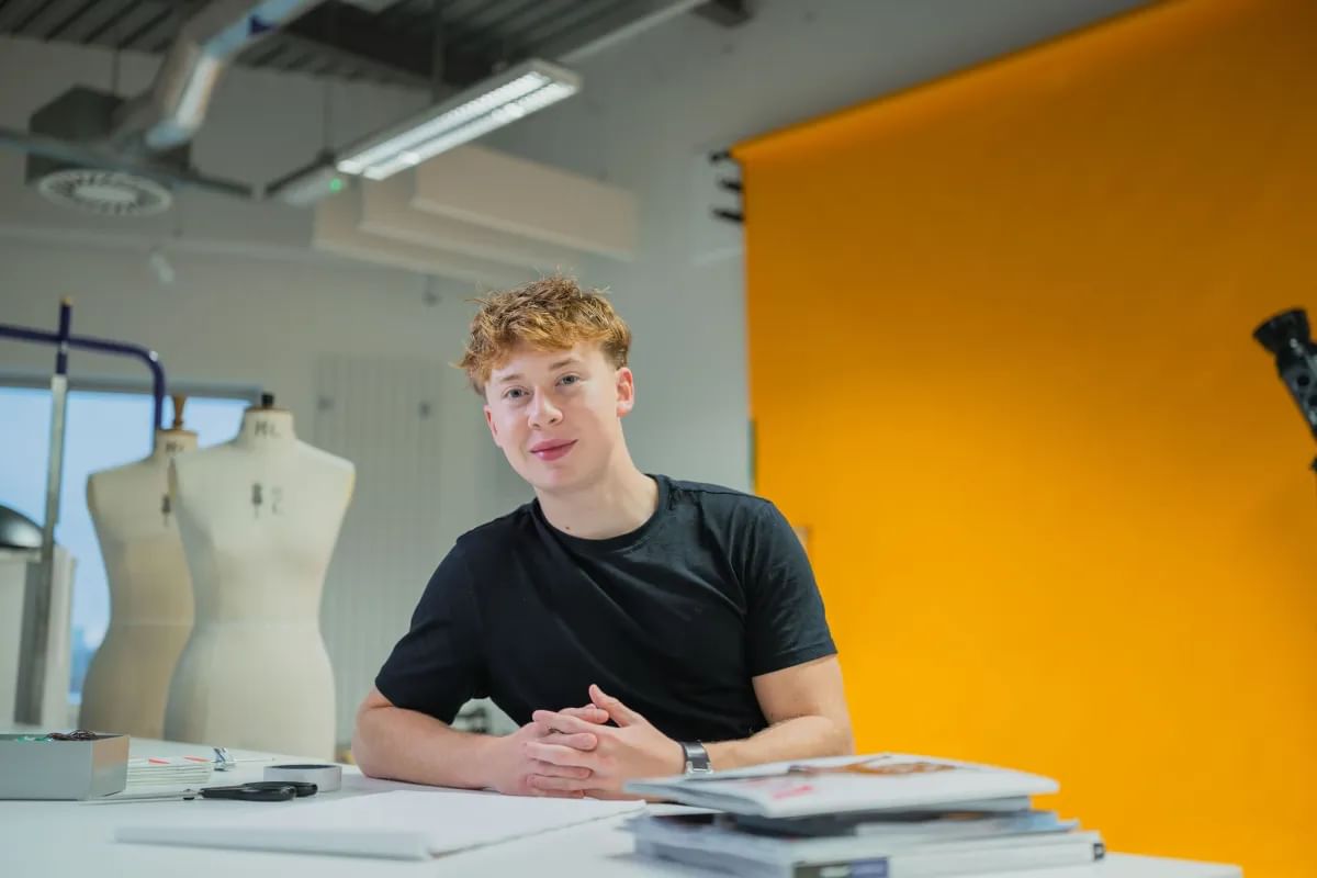 Dylan sits at a desk, dressed in stylish attire, with a bright yellow wall in the background.