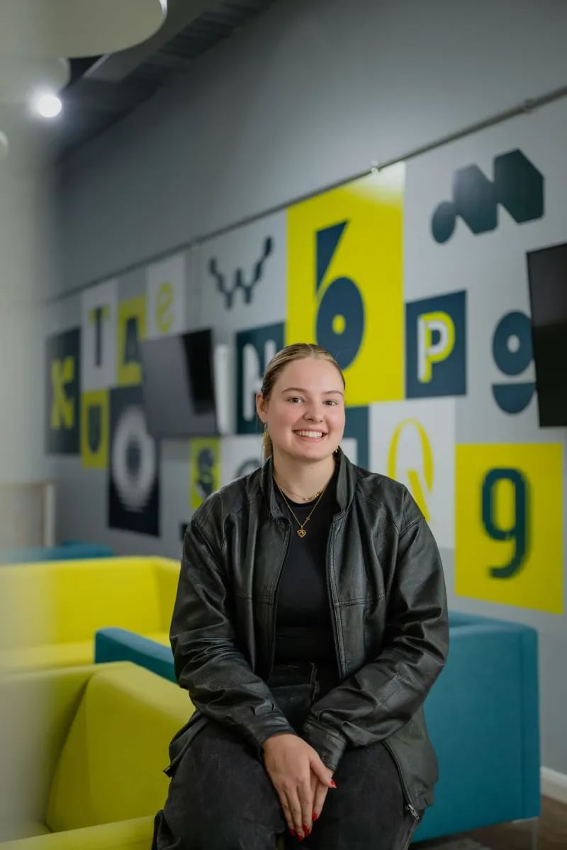 Graphic Communications student smiling at camera. She is wearing a black leather jacket and there are green chairs in the background.
