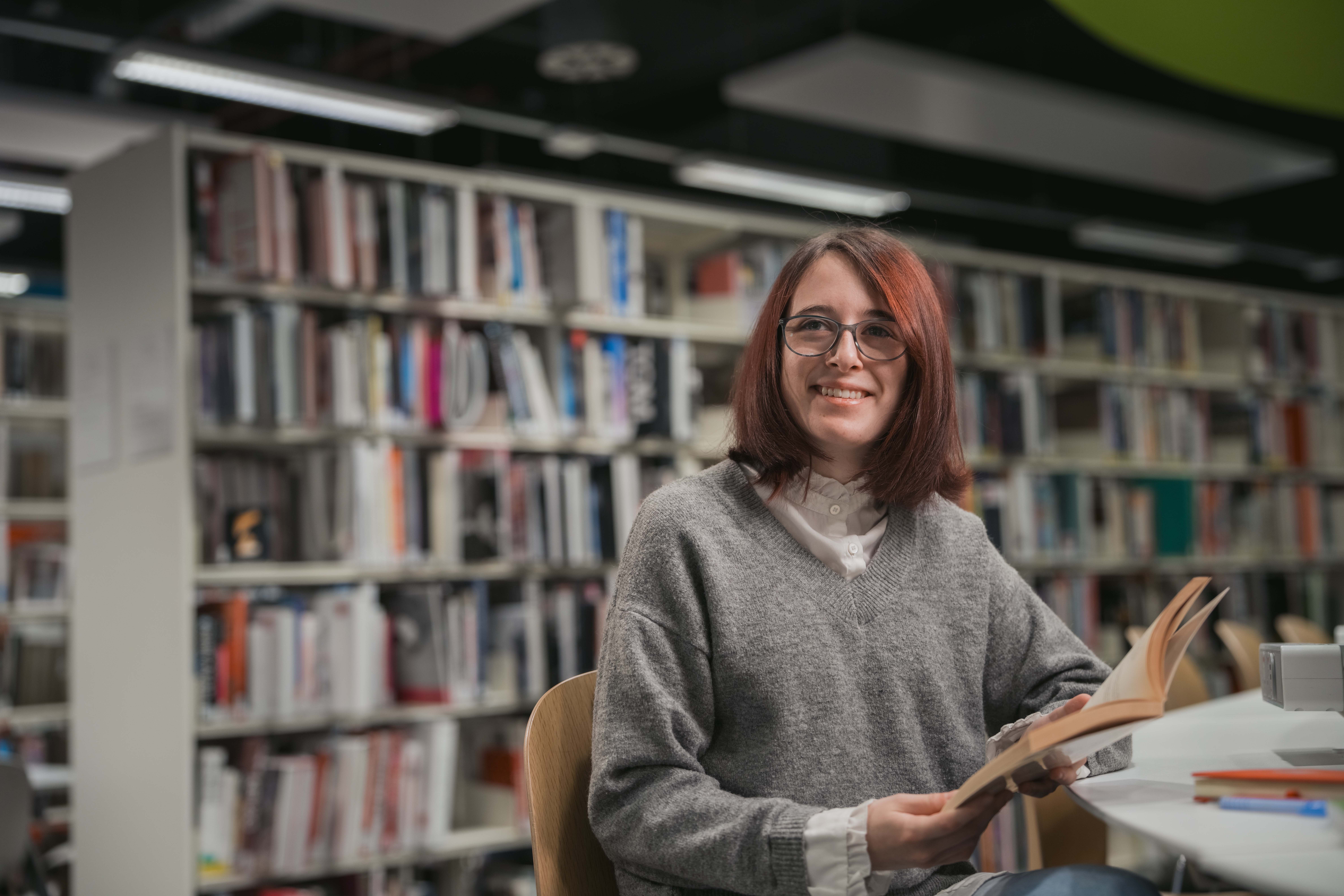 Smiling history student seated in a library, holding an open book at a table, profile shot with bookshelves in the background.