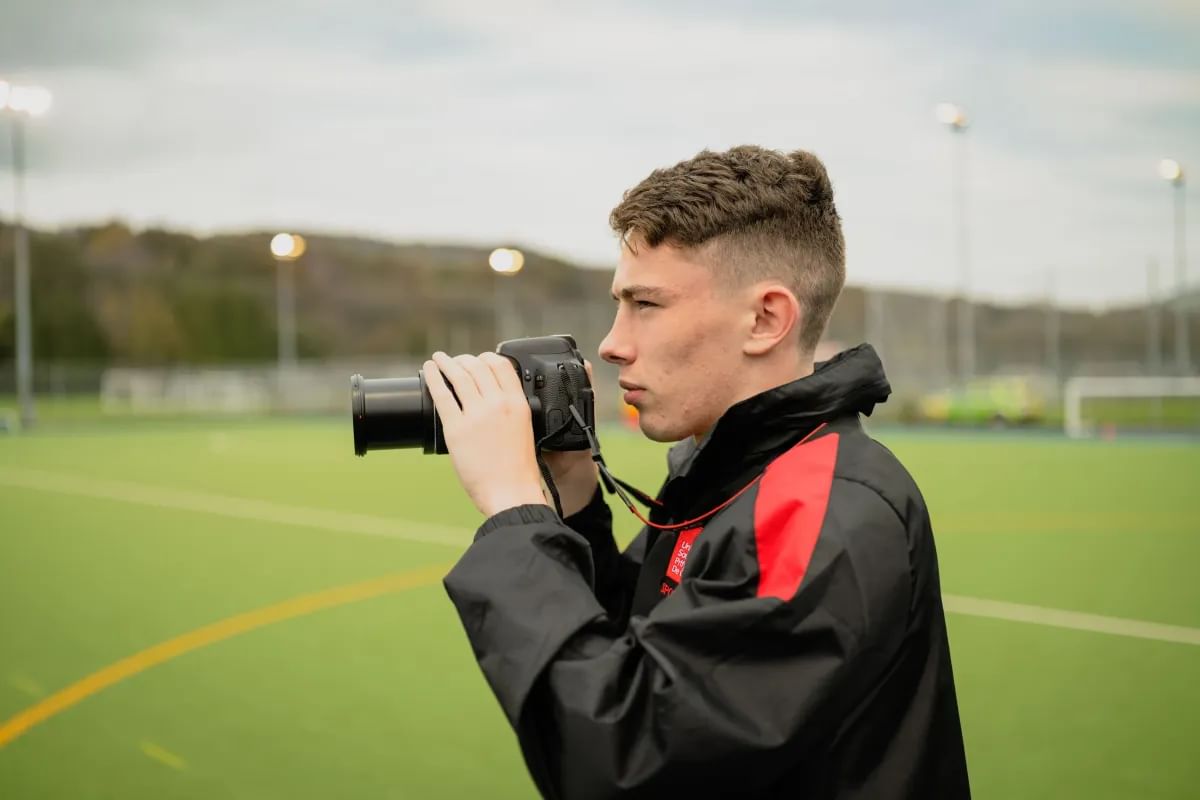 A student in sports journalism photographs a football match with his camera on the pitch
