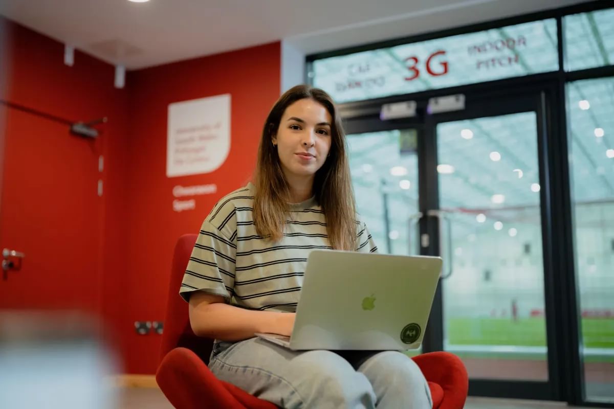 Sports Journalism student, Leah, poses for a photo with a laptop in front of her. She is sat in a room overlooking the football pitch