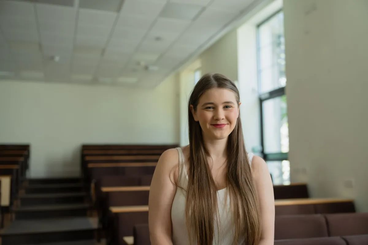 Shayla smiles at camera with lecture hall seats behind her. She has long dark hair and is wearing a white vest