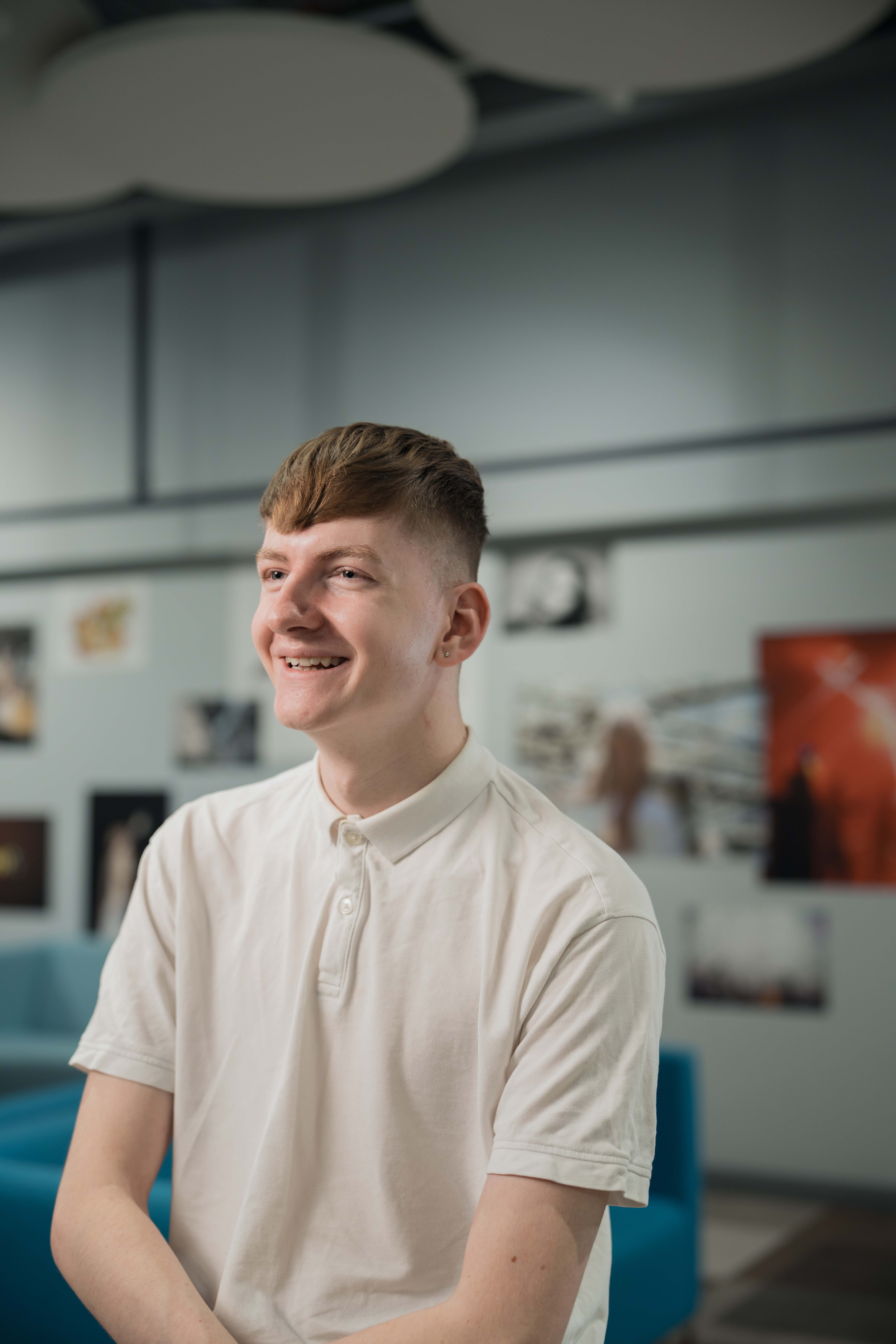 Marketing student, Sam, sits in front of colourful artwork whilst smiling at the camera