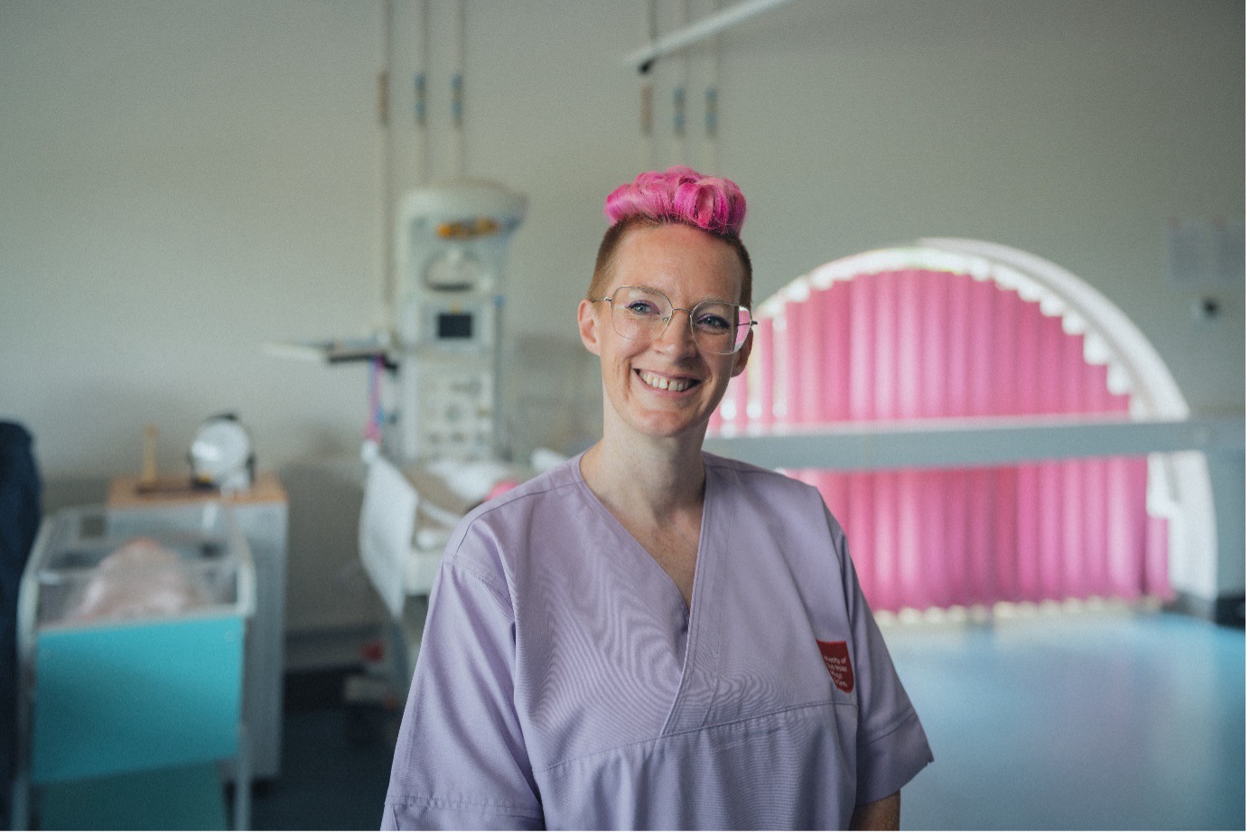 Midwifery student Kara smiles at camera, wearing purple scrubs in the clinical simulation centre