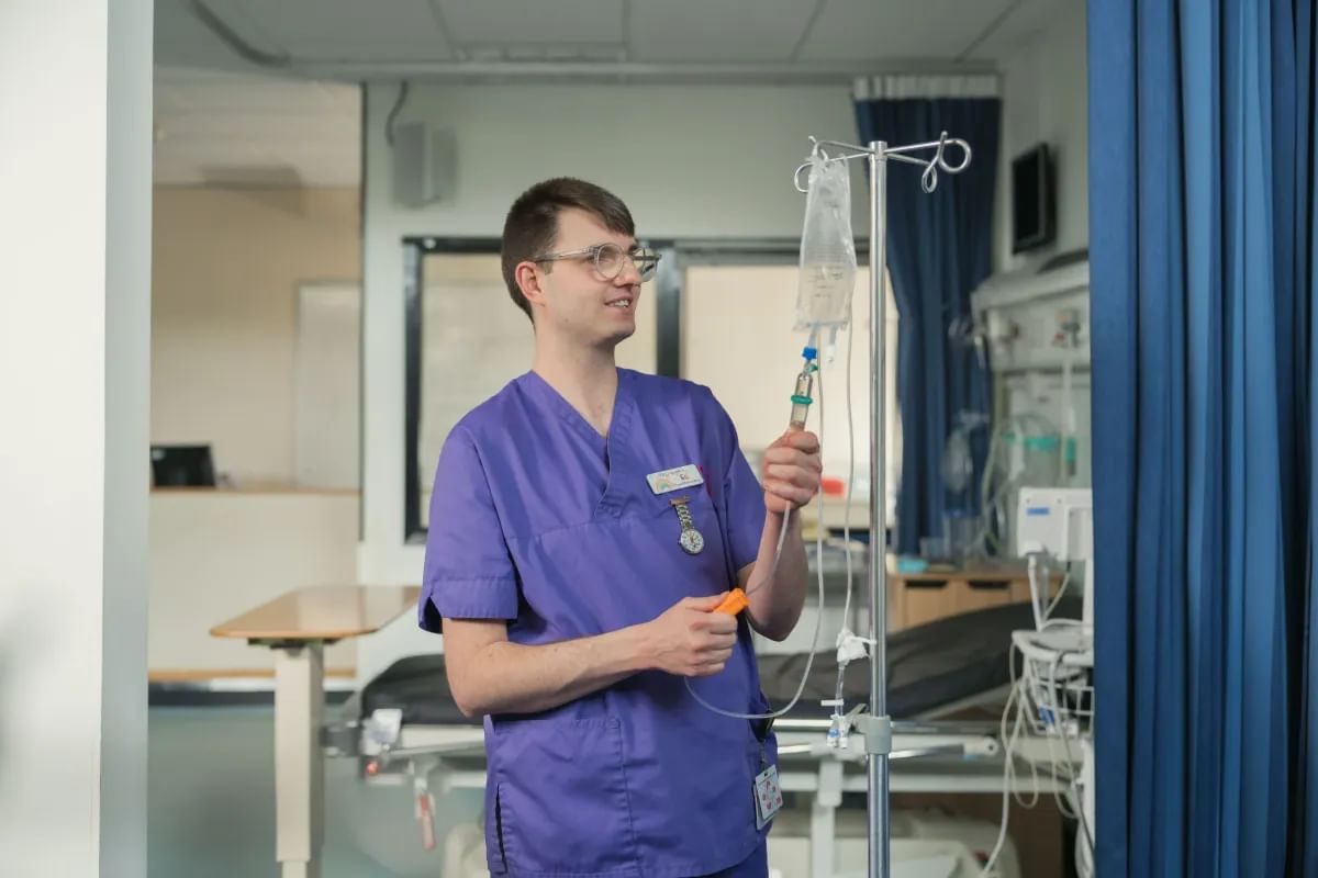 A male nurse in purple scrubs adjusts an IV drip in a hospital room. He is wearing glasses and a name badge, and is smiling while holding the IV tubing. Behind him are medical equipment, a hospital bed, and blue privacy curtains.