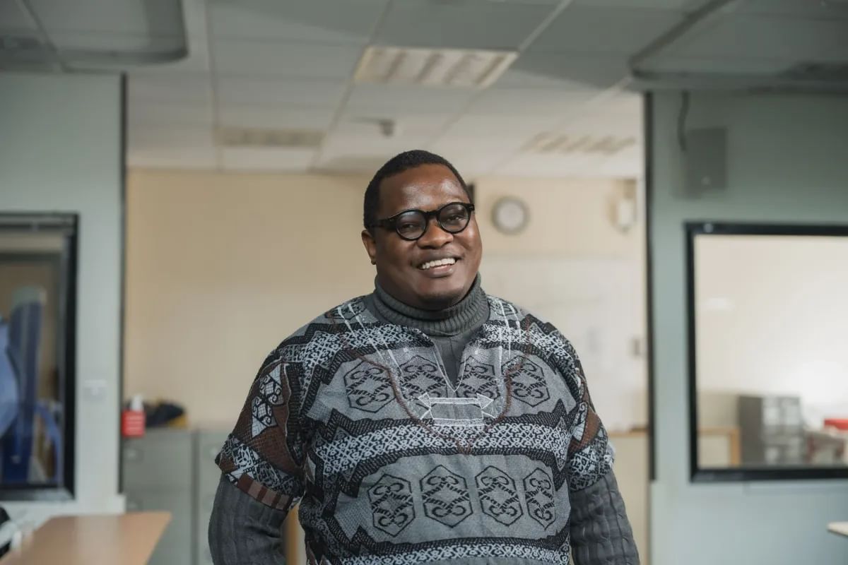 A man wearing glasses and a patterned sweater over a turtleneck stands smiling in a well-lit room with desks and windows.