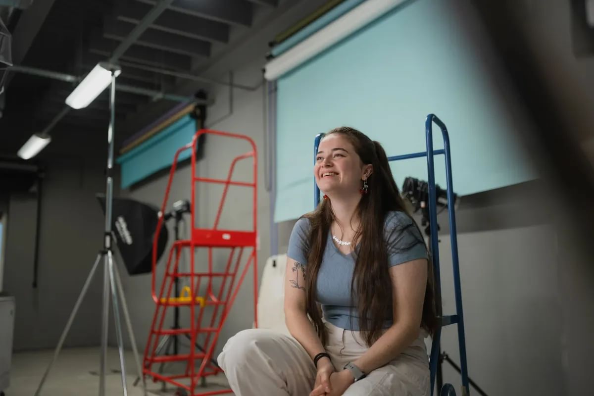 Ailish in casual clothes sitting on a chair, posing for the camera in a bright, photography studio.
