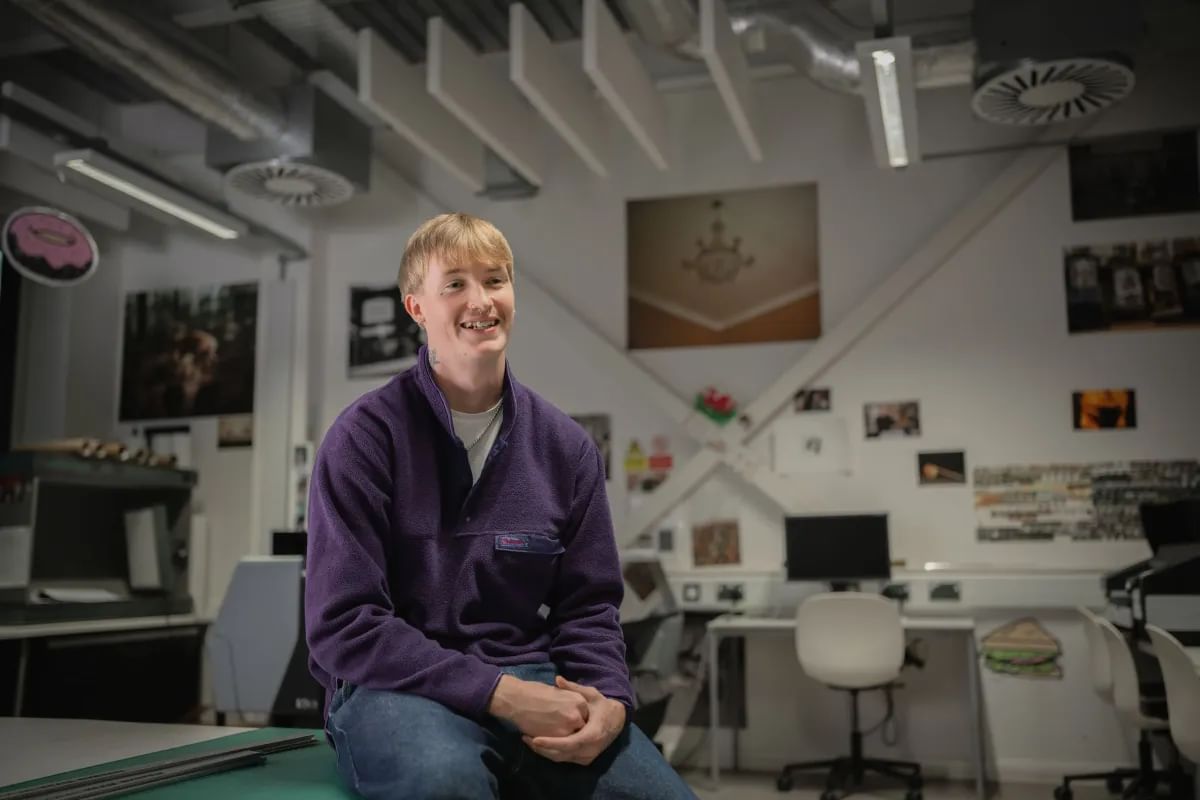 Photography student smiling for a photo, sitting on a desk in a classroom