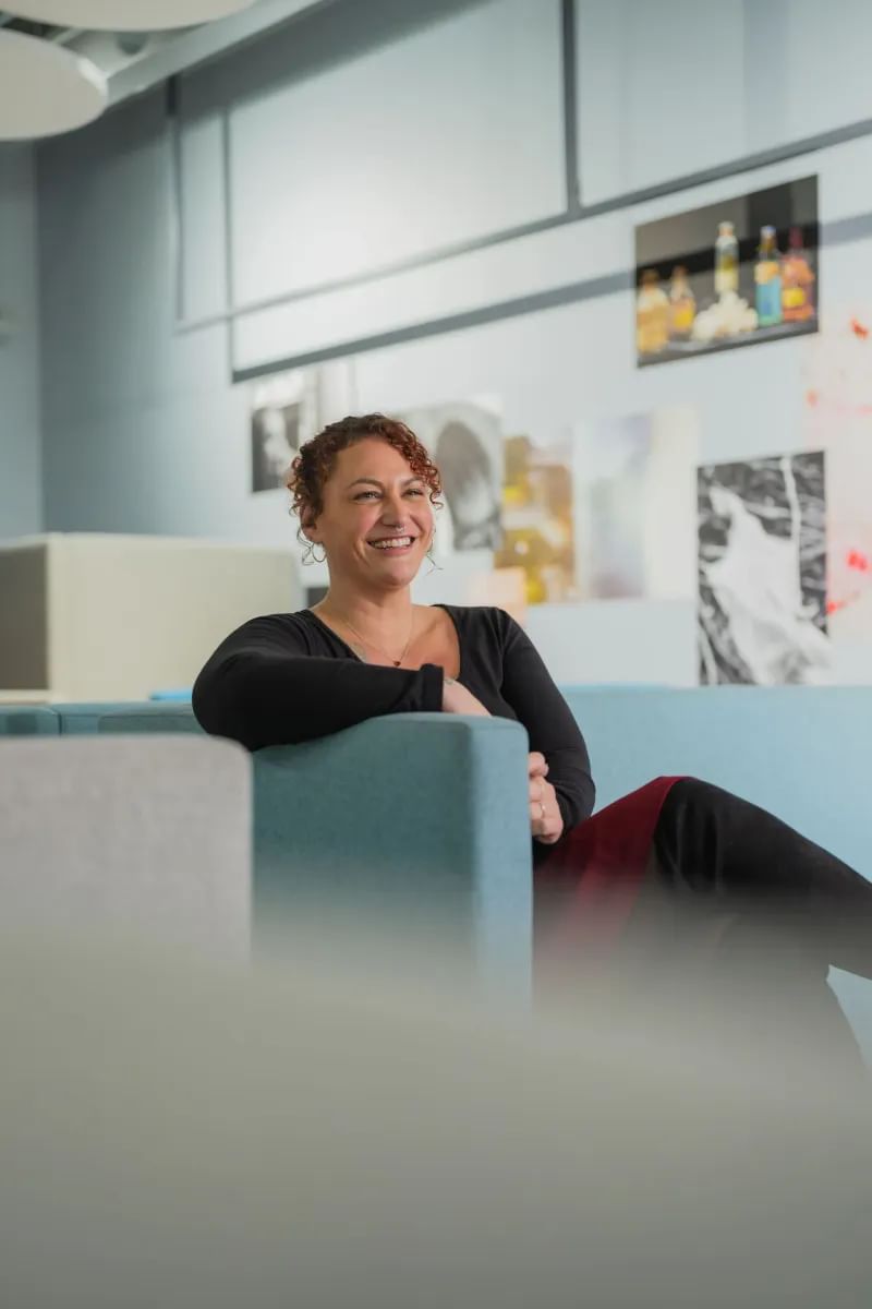 Photography student smiling for a photo, sitting on a sofa surrounded by a wall full of photographs