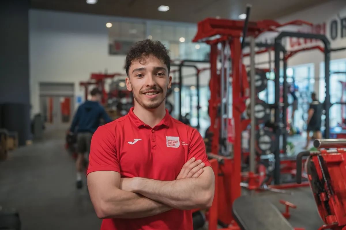 A sport student in a red polo shirt stands in a strength and conditioning gym, captured in a profile shot.