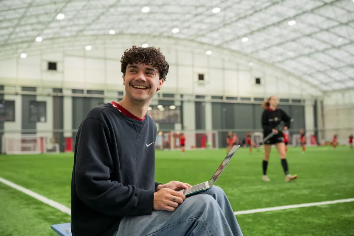 Sports Journalism student, Freddie, looks off to the side whilst sat on the side of a football pitch