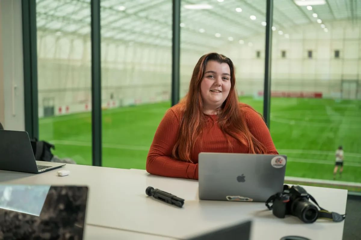 Sports Journalism student, Seren, poses for a photo with a laptop in front of her. She is sat in a room overlooking the football pitch