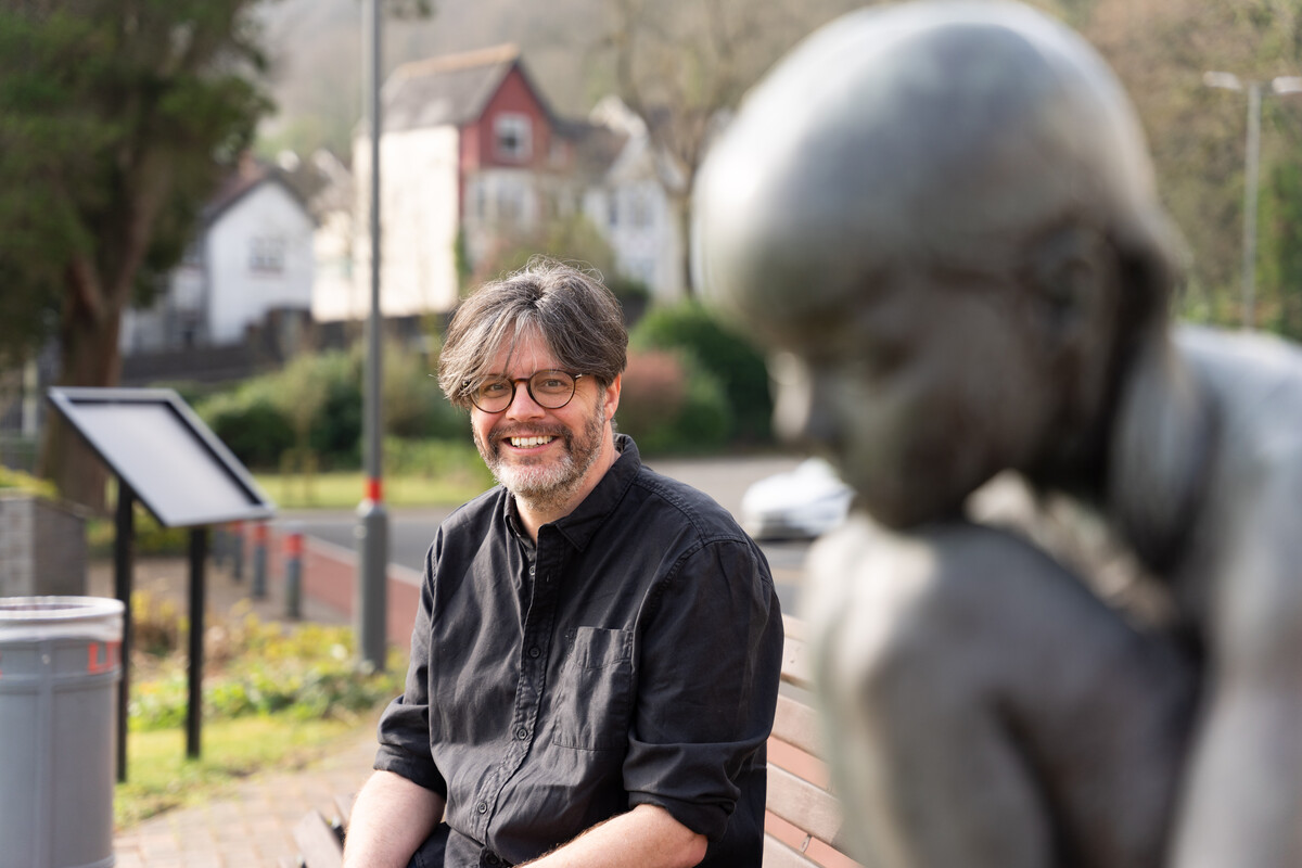 Dr Jonathan Durrant smiles at the camera while sat on a bench near a statue outside the Crawshay building on Treforest