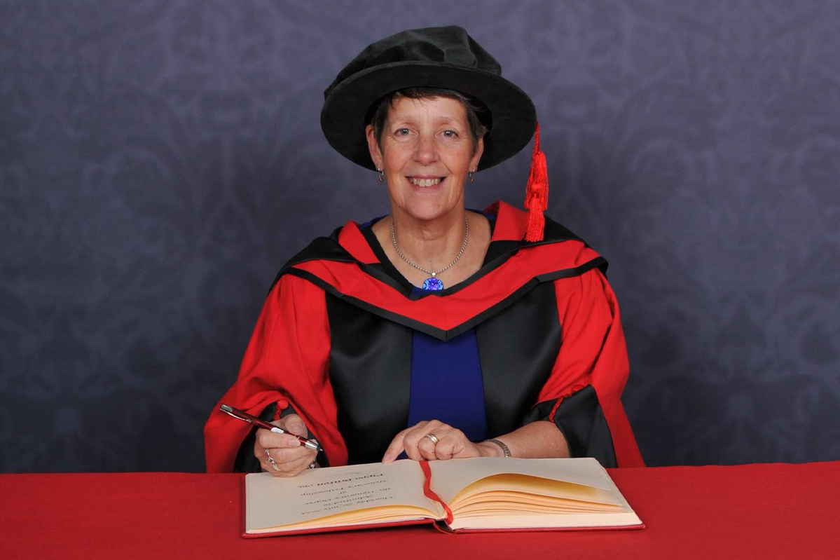 Pippa Britton writes in a large book on a red desk and smiles at the camera while wearing a doctorate's cap and robe in front of a blue backdrop