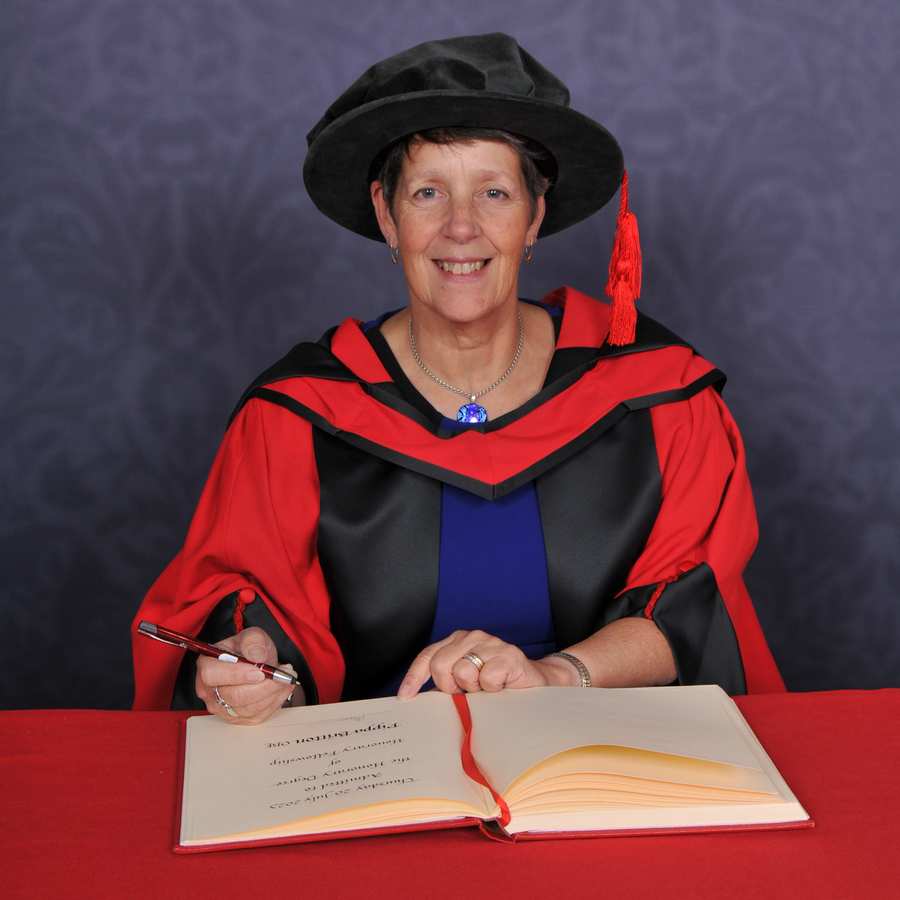Honorary fellow Pippa Britton smiles at the camera as they write in a large book on a red desk while wearing a PhD cap and gown