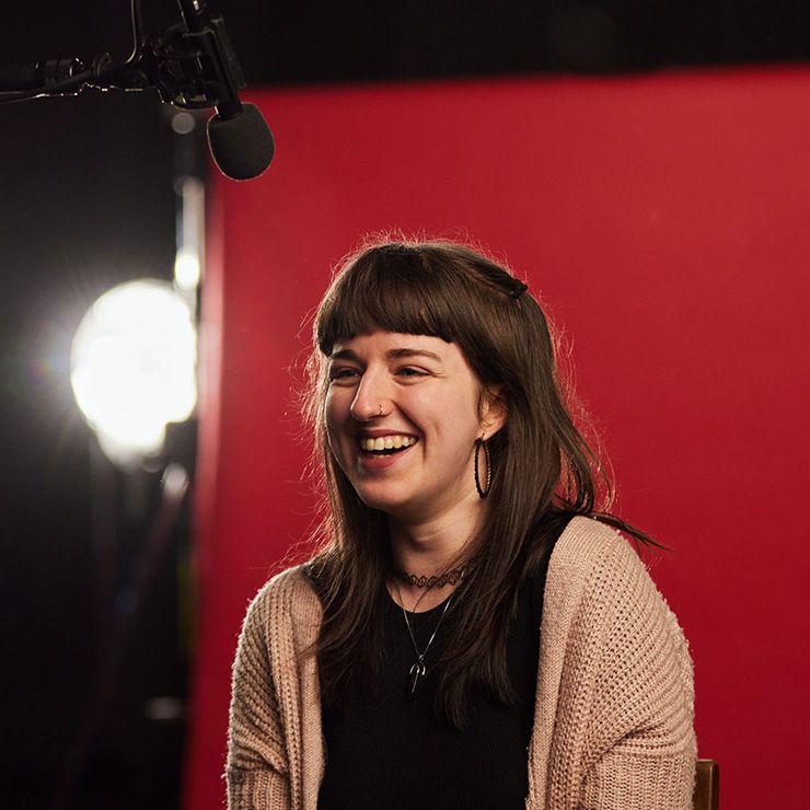 A student laughing in front of a red backdrop with filming lights in the background.